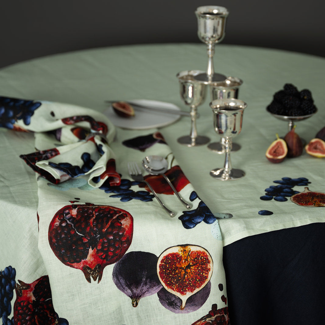Table setting with a decorative tablecloth featuring pomegranates and figs, accompanied by silver goblets and fruit.